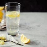 Homemade refreshing infused detox water or lemonade in glass with grated ginger and lemon on grey background close-up with copy space for text. Healthy vegan drink concept. Selective focus