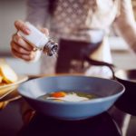 Close up of caucasian woman adding salt in sunny side up eggs while standing in kitchen next to stove.