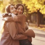 Side profile of young pretty smiling brunette twin girls hugging and having fun in casual coat standing close to each other at autumn sunny park alley on blurry background